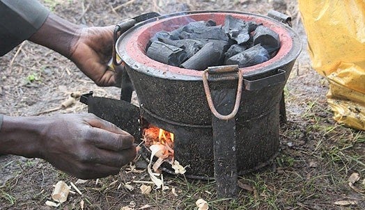 This image shows hands lighting the bottom part of a circular stove, with charcoal in a bowl-like container on top. The charcoal is smoking. The stove is sitting on the ground. It is about the size of a soccer ball.