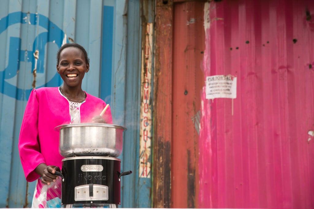 This image shows a young woman stirring food in a metal bowl that rests on a small metal appliance similar in size to the Jiko. Smoke is rising from the bowl. 