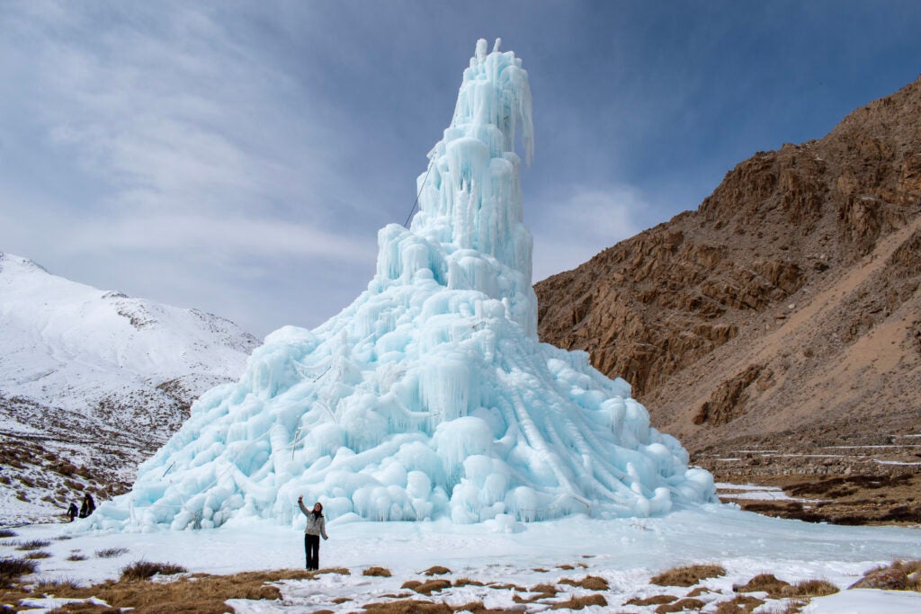 A woman stands in front of a large ice tower in the arid mountains of the Himalayas to provide scalar perspective on the size of the stupa.