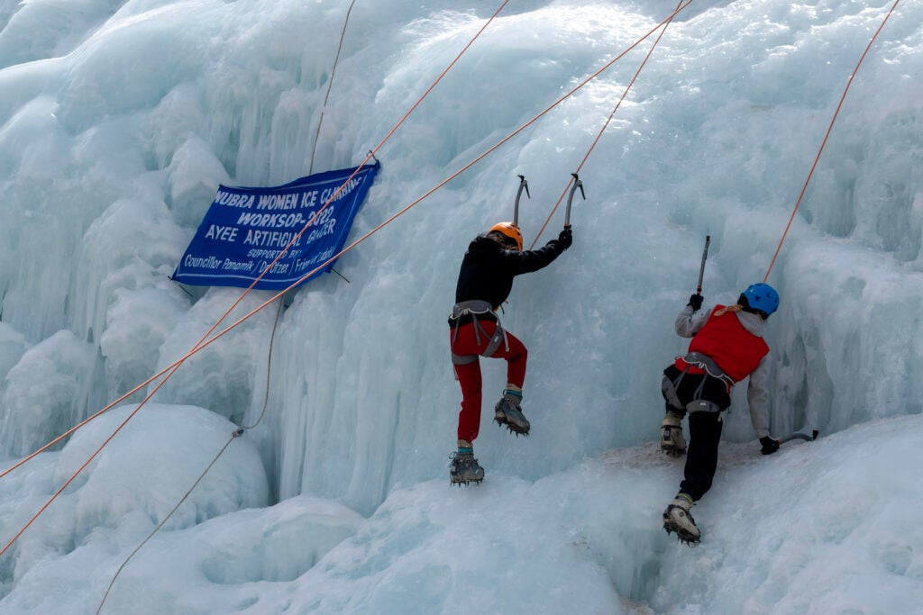 Two women with ice axes climb up an ice stupa. 