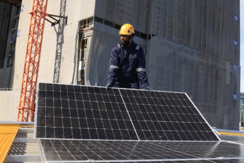 Male technicians carrying photovoltaic solar module on roof top of factory. Engineers in helmets installing solar panel system outdoors. Concept of alternative and renewable energy.